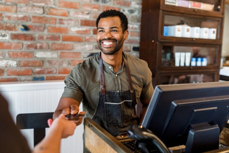 A smiling small business owner wearing a dark apron and olive green shirt stands behind a counter, reaching out to accept a customer’s credit card or phone for payment. The setting is a stylish shop with a rustic brick wall and wooden shelving stocked with products. A point-of-sale terminal and barcode scanner are visible on the counter. The overall atmosphere is warm, welcoming, and professional, conveying friendly customer service and modern retail operations.