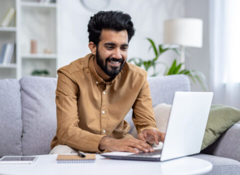 A young man working from his living room uses a laptop to send payments for his small business.