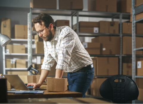 small business owner in a warehouse using a laptop to check his payments platform is PCI-compliant.
