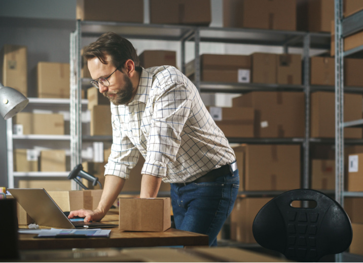 small business owner in a warehouse using a laptop to check his payments platform is PCI-compliant.