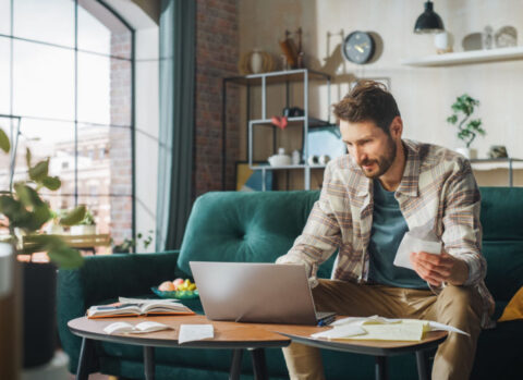 A small business owner sitting on a sofa in his living room and filing his taxes online using a laptop.