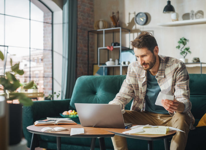 A small business owner sitting on a sofa in his living room and filing his taxes online using a laptop.