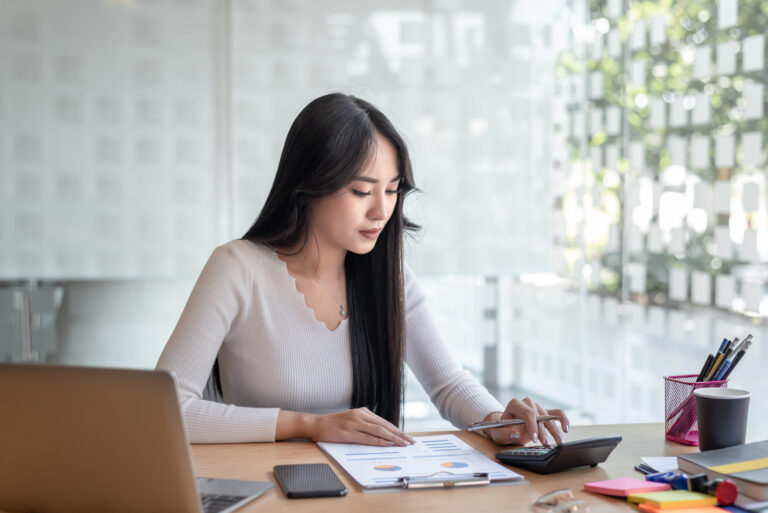 Woman working at a desk in a modern office space, focused on financial documents. She uses a calculator while reviewing charts and graphs on a printed report. A laptop, smartphone, pens, sticky notes, and a coffee cup are also on the desk. Sunlight filters through patterned glass walls in the background.