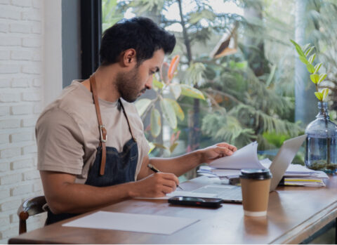 A coffee shop owner using a laptop and calculator to manage B2B payments to vendors and suppliers.