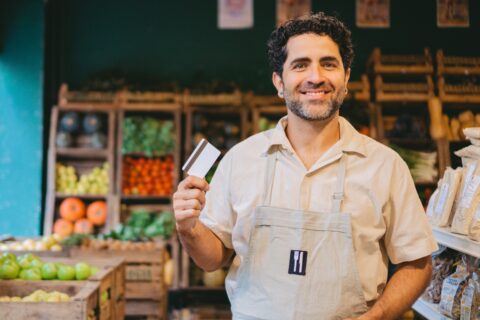 Smiling male shopkeeper in a beige shirt and apron holding a credit card inside a small grocery store or market. Fresh produce like tomatoes, apples, and squash are displayed on wooden shelves in the background.