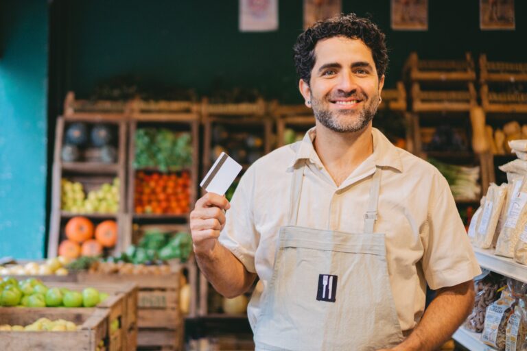 Smiling male shopkeeper in a beige shirt and apron holding a credit card inside a small grocery store or market. Fresh produce like tomatoes, apples, and squash are displayed on wooden shelves in the background.