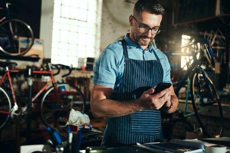 A man wearing glasses and a striped apron stands in a bicycle repair shop, looking down at a smartphone in his hands. He appears focused and calm, possibly checking orders or managing payments. The workshop is filled with tools, bike parts, and several bicycles in the background, giving a warm, industrious atmosphere with sunlight streaming through large windows.