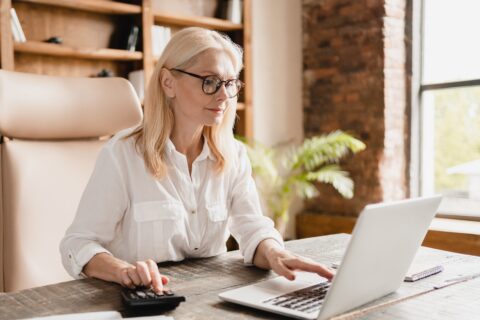 A woman with blonde hair and glasses, wearing a white button-down shirt, working at a rustic wooden desk. She is using a laptop with one hand and a calculator with the other. The background shows a cozy office space with bookshelves, a plant, and a brick wall.