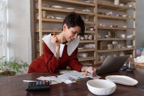 Small business owner managing expenses at a pottery studio, reviewing receipts with a laptop, calculator, and handmade ceramics on the table.