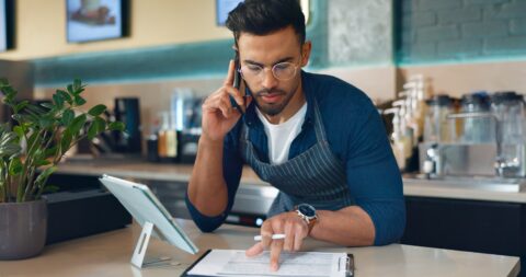 Small business owner in a café wearing an apron and glasses, multitasking by speaking on the phone while reviewing documents on a clipboard and using a tablet at the counter, representing modern entrepreneurship and business management.