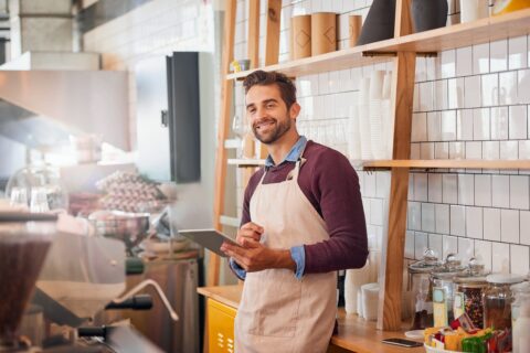 Smiling small business owner wearing an apron stands behind the counter of a cozy coffee shop, holding a tablet. The shop interior features white tiled walls, wooden shelves with cups and jars, and a welcoming, organized atmosphere