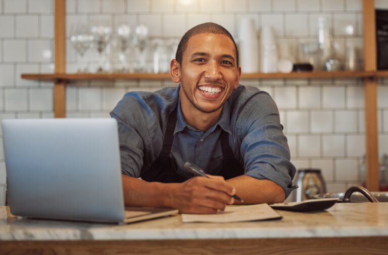 Smiling small business owner wearing an apron, sitting at a counter with a laptop, writing on a document.