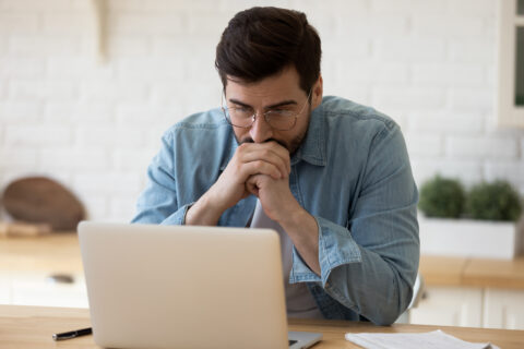 Focused young man in glasses working on laptop at home office desk, looking stressed or thoughtful.