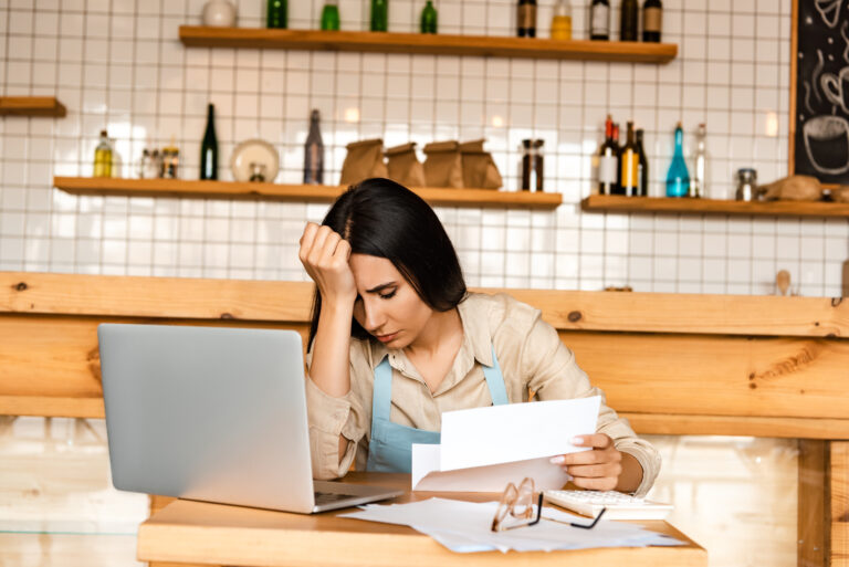 Frustrated woman in an apron sitting at a desk with a laptop and papers, holding her head in one hand.