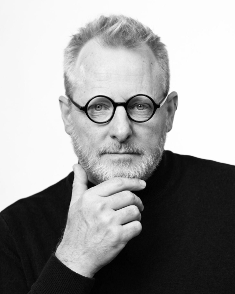 A black-and-white portrait of Jason Blumer, a man with a graying beard and round black glasses, wearing a dark turtleneck. He is posed with a hand under his chin against a white background, looking directly at the camera with a thoughtful expression.