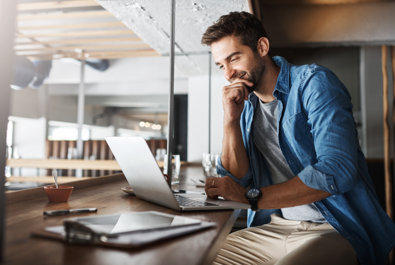 A man in a blue shirt works on a laptop at a wooden table in a sunlit, modern co-working space.