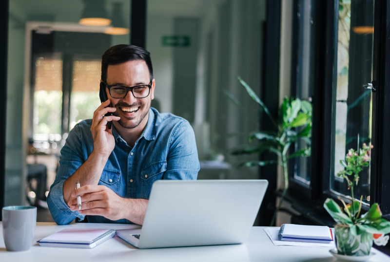 A man with glasses sits at a desk, laughing while on a phone call in a bright office.