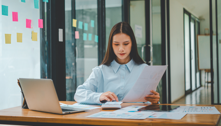 Business professional reviewing financial reports and calculating data at a desk with a laptop and paperwork, representing accounting, bookkeeping, financial analysis, or small business finance management.