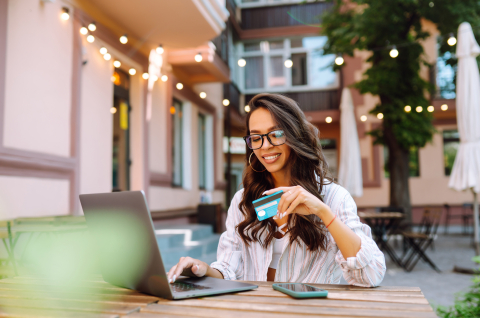 A smiling woman wearing glasses and a striped shirt sits at an outdoor cafe, holding a credit card while using a laptop. Warm string lights and a patio setting are visible in the blurred background.