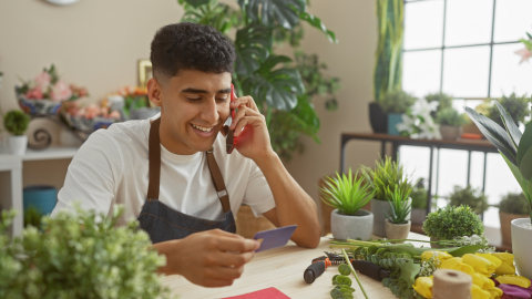 A smiling young man wearing a florist's apron sits at a wooden table in a plant-filled shop. He is talking on a red smartphone while holding a blue credit card, surrounded by fresh flowers and potted plants.