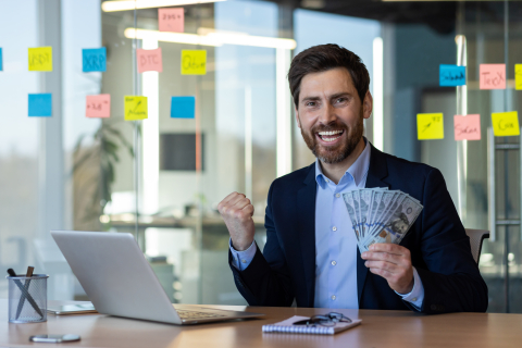 Business owner celebrating positive cash flow and financial success, holding cash at desk in modern office with financial planning materials