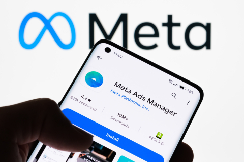 A person’s hand holds a smartphone displaying the Meta Ads Manager app download page in front of a large, glowing Meta logo.
