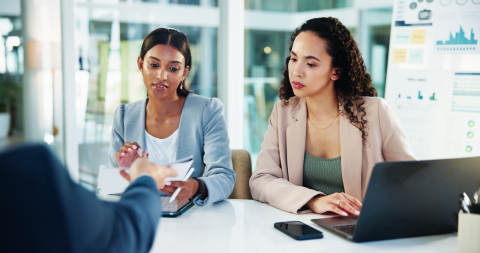 Two professional women sitting at a desk during a business meeting, looking focused as they review documents being presented by a colleague.