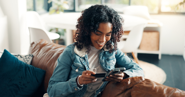 A smiling young woman with curly hair sits on a brown leather sofa, holding a credit card and using her smartphone to make an online purchase.