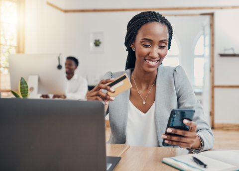 A smiling professional woman in an office setting holding a credit card while using her smartphone, with a laptop and a colleague in the background.