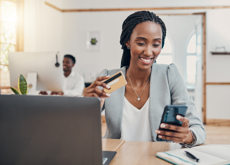 A smiling professional woman in an office setting holding a credit card while using her smartphone, with a laptop and a colleague in the background.