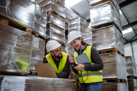 Two warehouse workers wearing hard hats and safety vests smile while looking at a laptop together amidst tall stacks of shrink-wrapped pallets.